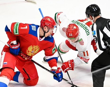 На изображении два хоккеиста в форме сборных России и Беларуси. Они борются за шайбу на льду, рядом находится судья. Игроки активно участвуют в игре, демонстрируя спортивное соревнование