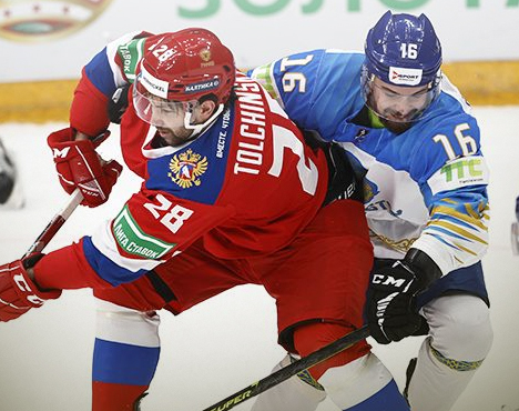 Хоккеист в красной форме сражается за шайбу с хоккеистом в бело-синей форме на льду. Оба игрока сосредоточены на игре, вокруг виден лёд и хоккейное снаряжение