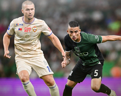 На изображении два футболиста на поле: один в светлой форме, другой в темной. Они сосредоточены на игре. На заднем плане размытые трибуны