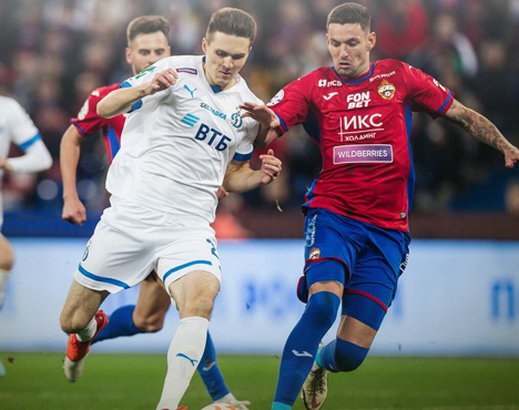 Футболисты двух команд в яростной борьбе за мяч на поле. Один игрок в белой форме, другой в красной. Они сосредоточены на игре, на фоне видны размытые фигуры болельщиков