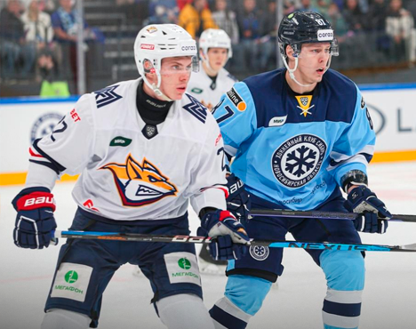 На изображении два хоккеиста в форме разных команд на льду во время матча. Один в форме бело-оранжевых цветов, другой — в голубо-синих. Они держат клюшки и сосредоточены на игре